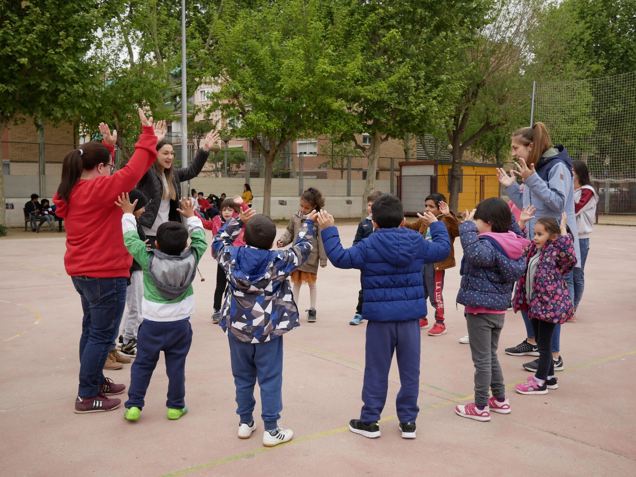 Nens i educadores en una rotllana a peus amb els braços a dalt, fent una activitat. Acció Comunitària als Barris