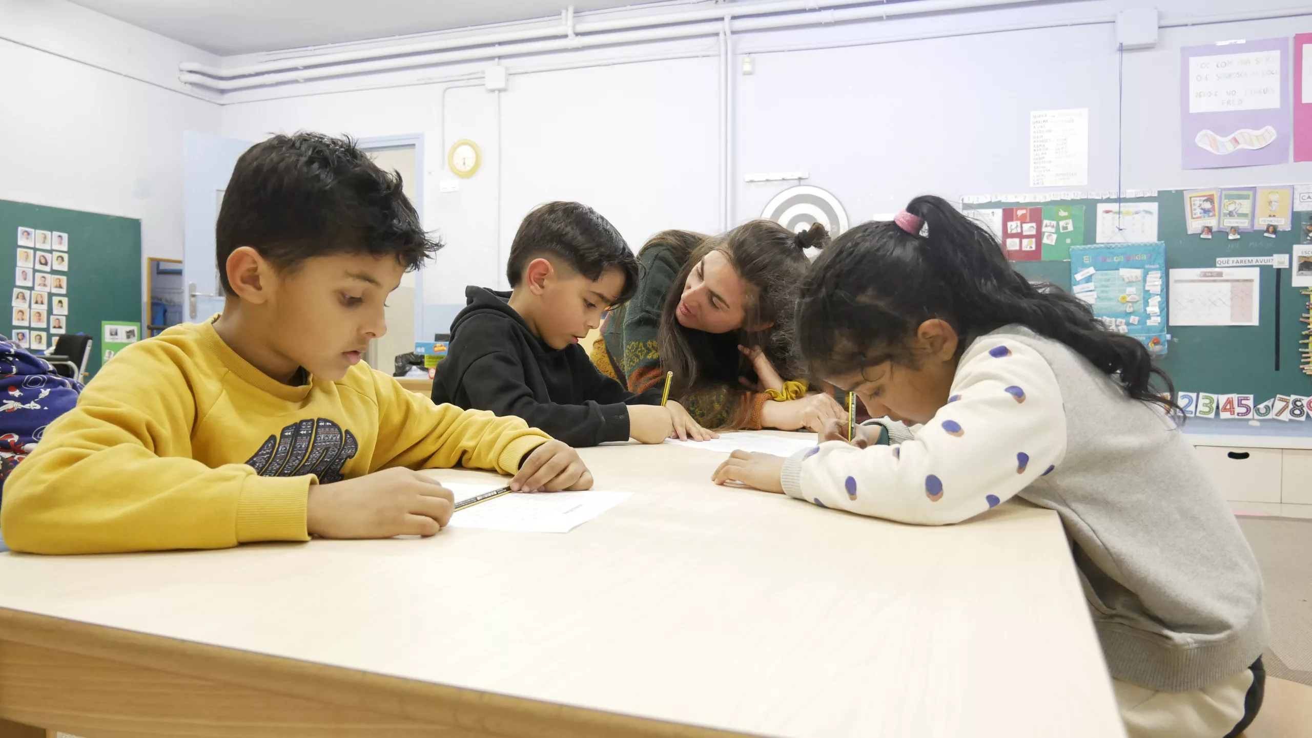 Tres infants seuen al voltant d'una taula fent un exercici escrit, amb una educadora social que els acompanya.