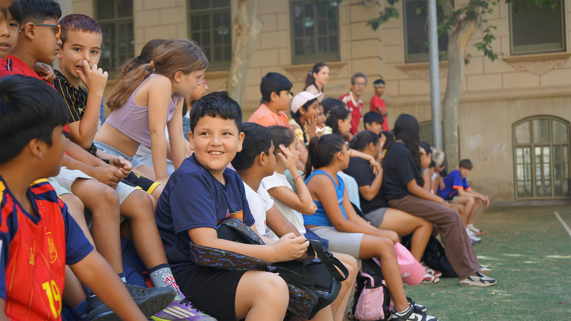 Nens i nenes asseguts un al costat de l'altre, en dos nivells diferents, al pati d'una escola.