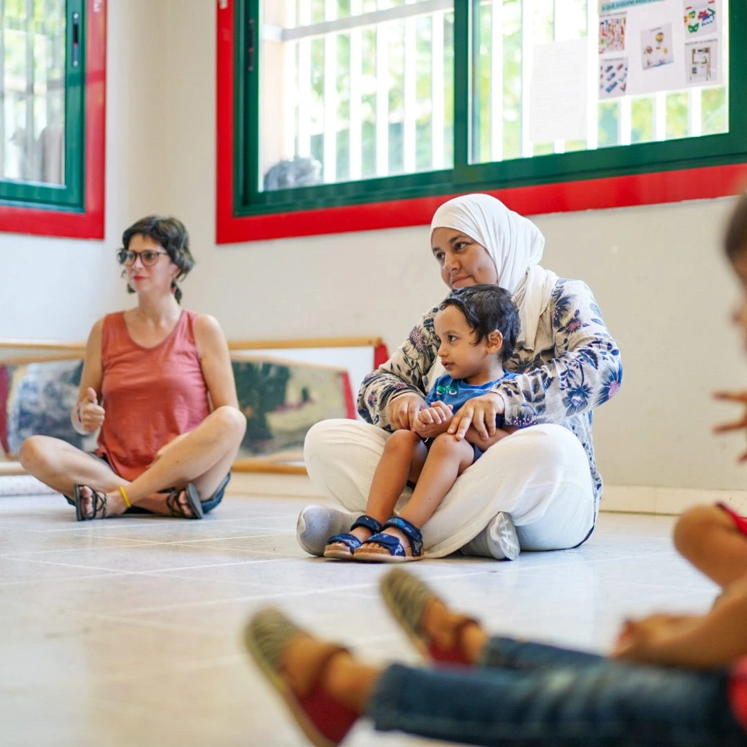 Una mare i el seu fill asseguts en el sòl junts amb una educadora del projecte +infància.