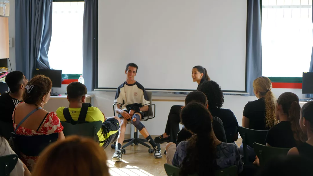 Aula amb joves asseguts en cadires d'esquenes, escolten la xerrada de l'Àlex Roca, amb la interpretació de la Mari Carme Maza.