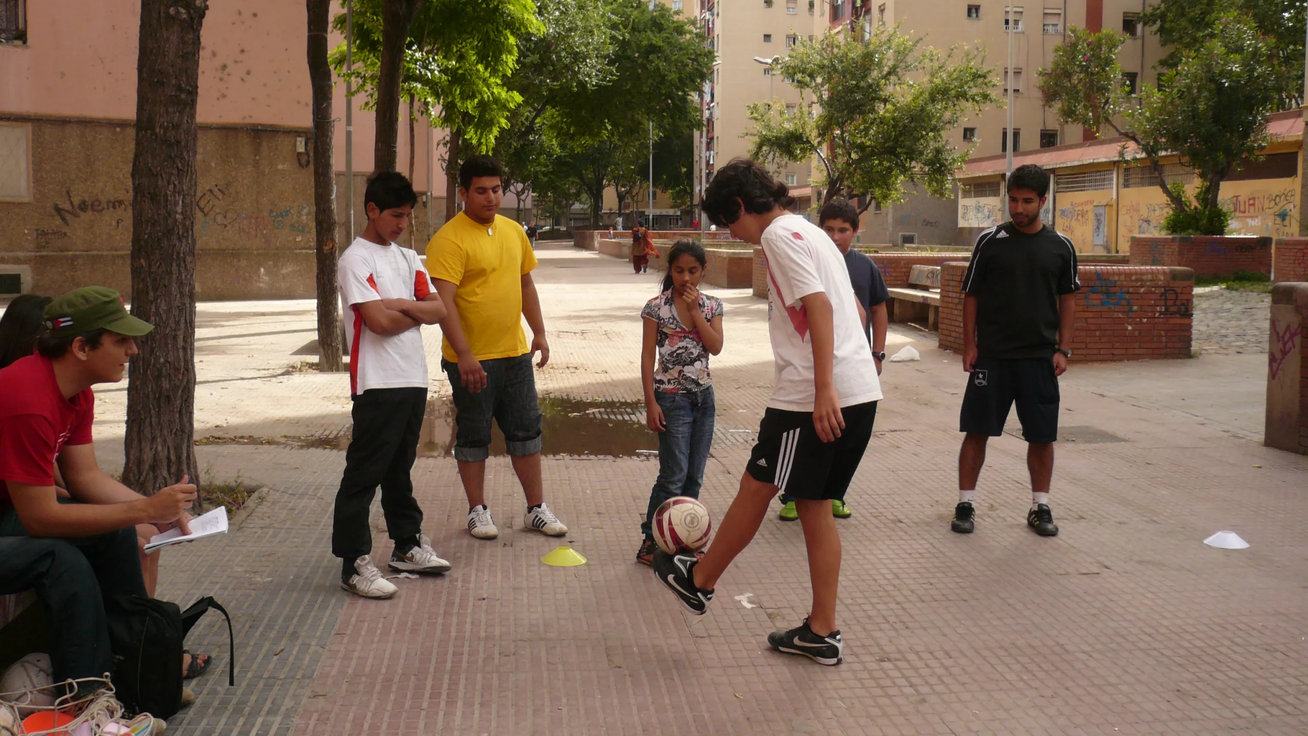 Noies i noies de peu miren com un altre noi juga amb una pilota amb el peu.