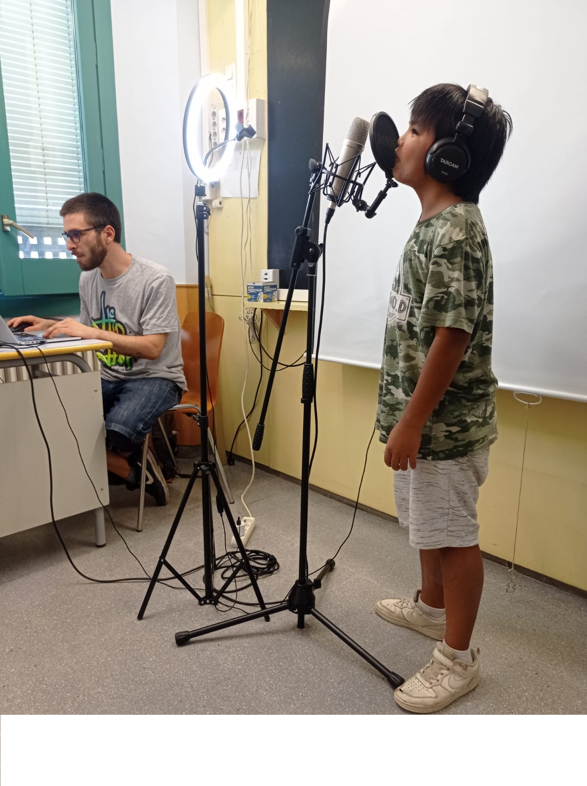 Nen de peu canta a un micro amb els auriculars posats, mentre un home amb un ordinador grava la veu.