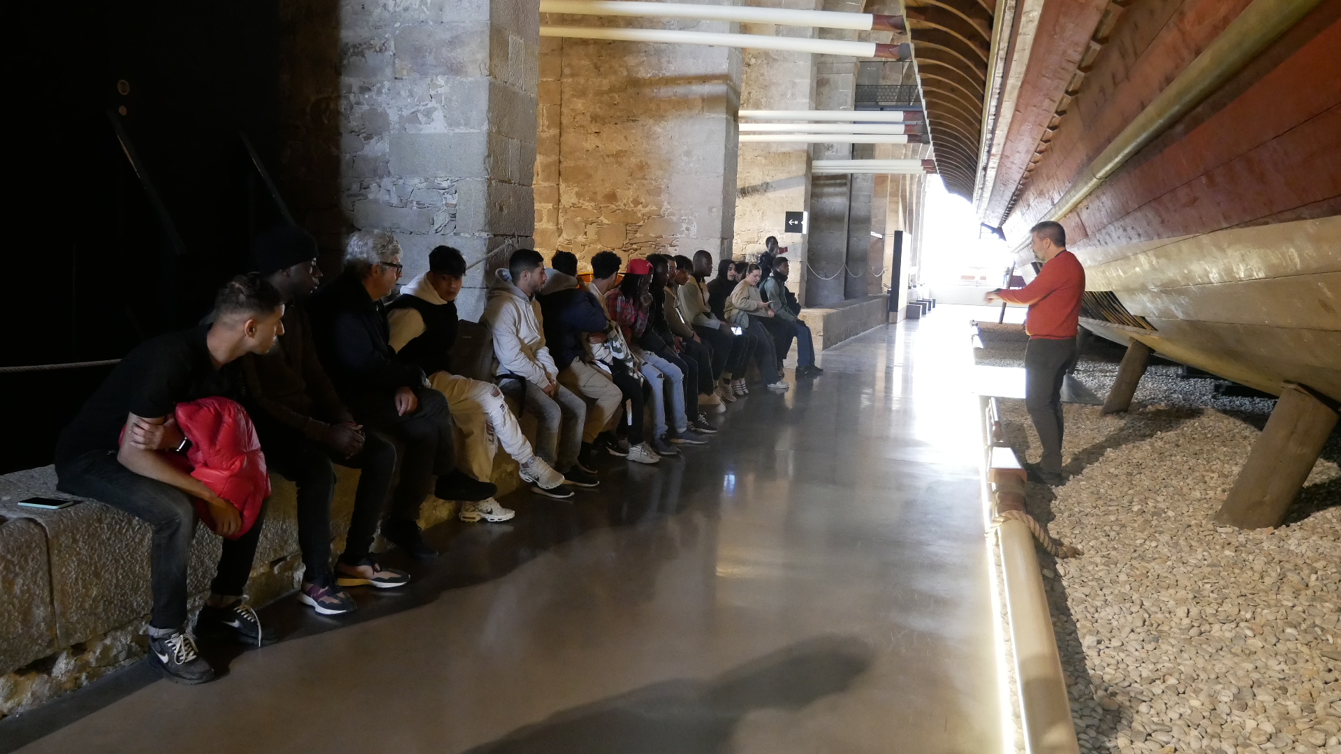 Nois asseguts escolten el guia d'una visita al Museu Marítim de Barcelona