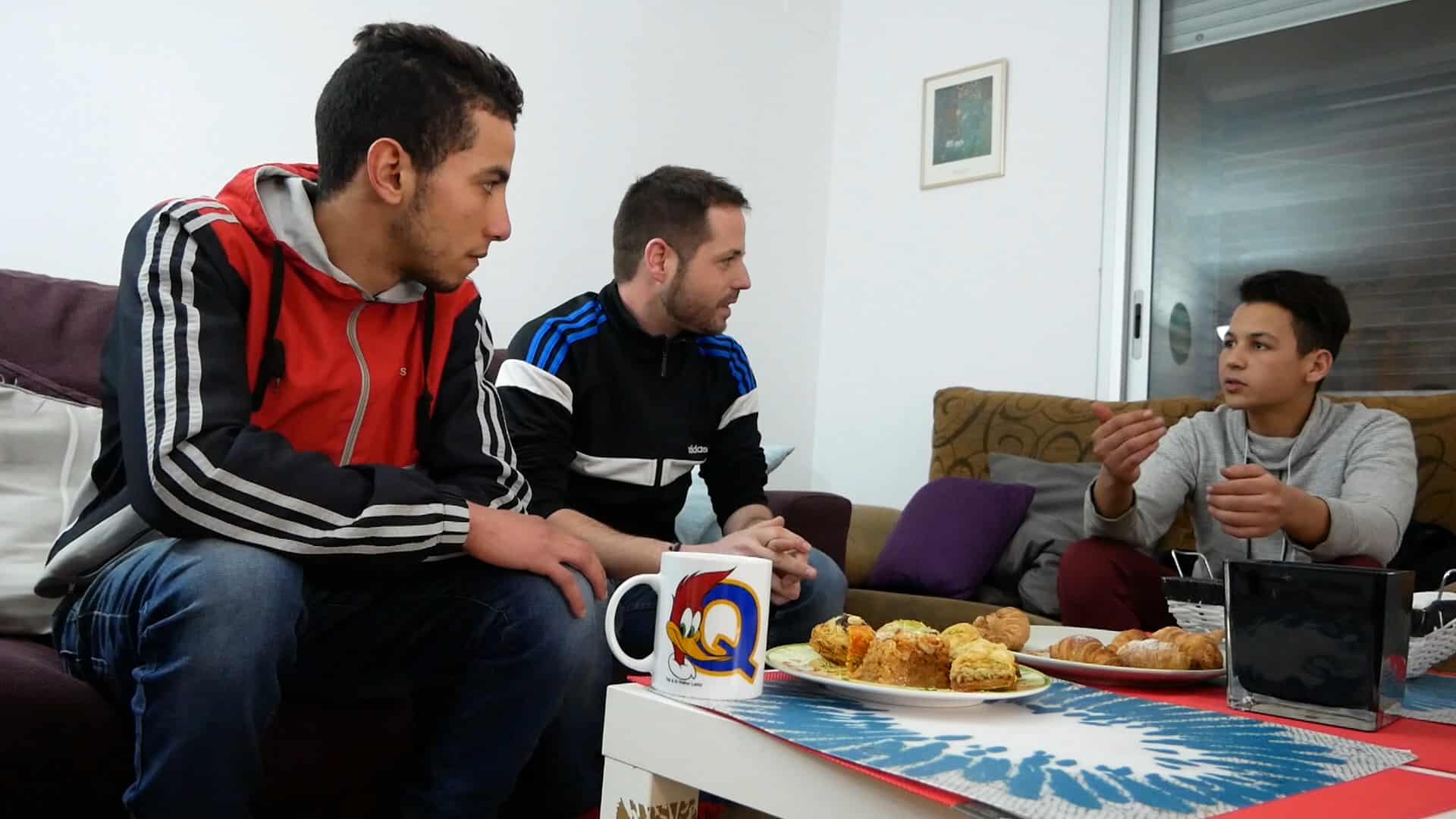 Tres nois asseguts en un sofà. Davant una taula amb pastes dolces i una tassa.